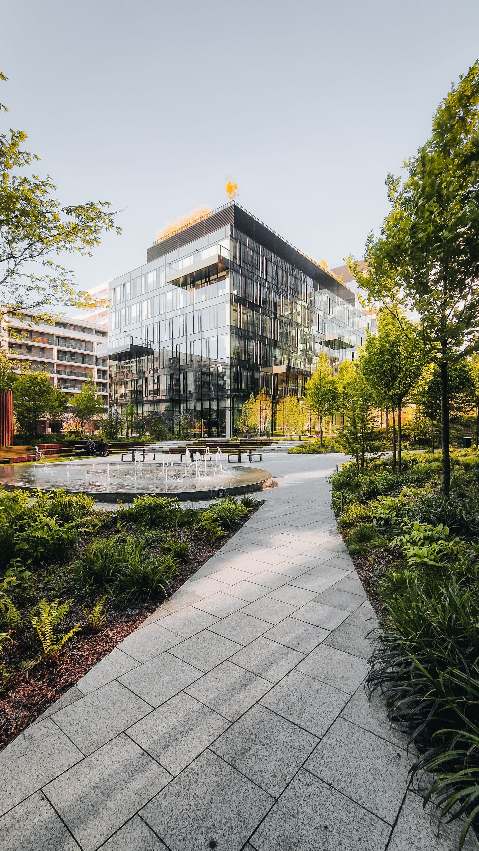 A sleek office building with glass facade surrounded by lush gardens and a central fountain.